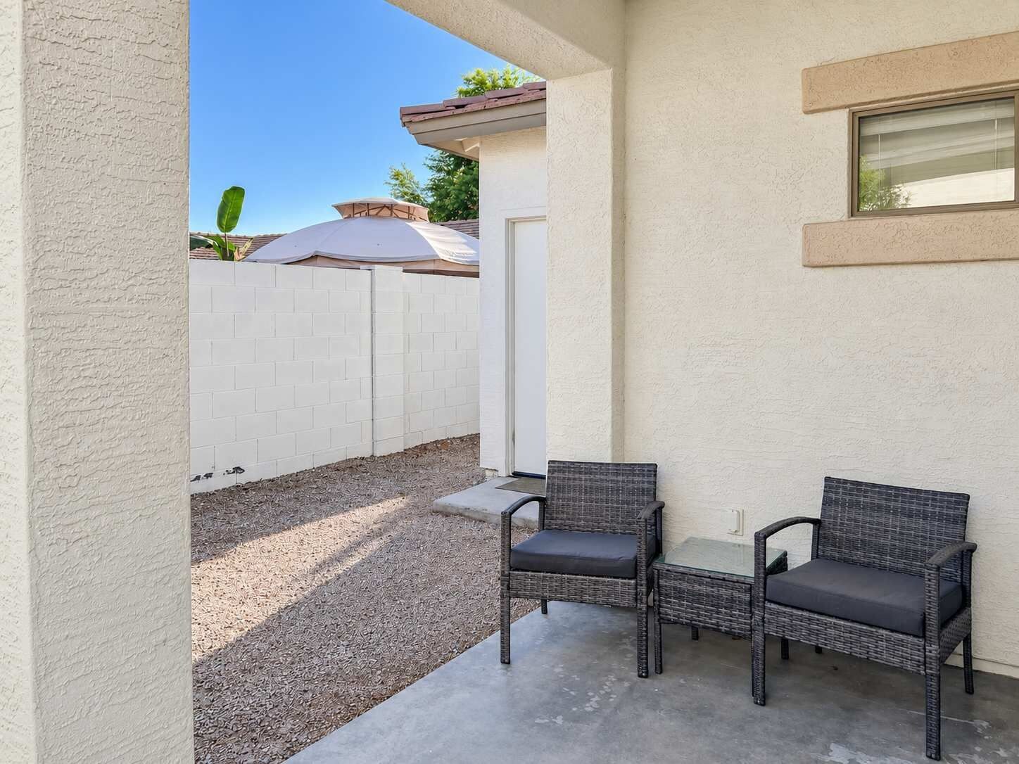 Ein Patio mit zwei Korbstühlen, einem kleinen Tisch, einem weißen Zaun und blauem Himmel.