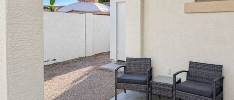 A patio area with two wicker chairs and a small table, a white fence, and a clear sky.