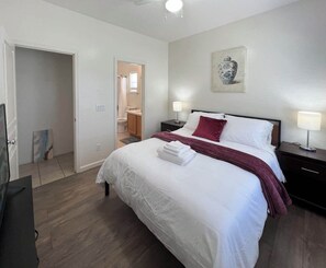 Ein Hotelzimmer mit einem großen Bett, einem Nachttisch mit Lampe, einem Gemälde an der Wand und einem durchgehenden Türspalte sichtbaren Badezimmer.