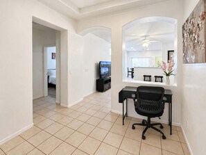 A modern living room with a black office chair, a flat-screen TV, and a dining area visible through an archway.