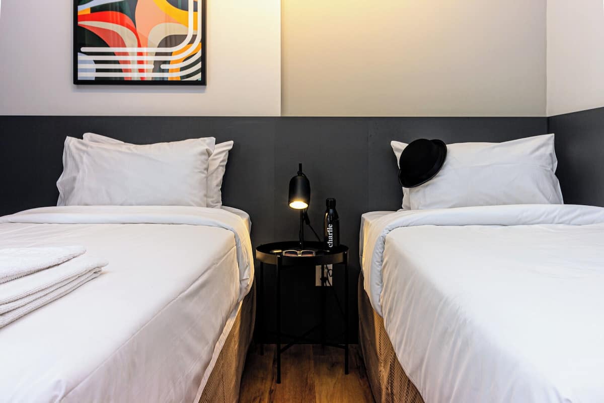 A hotel room with two beds, a small table with a lamp, and a colorful abstract painting on the wall.