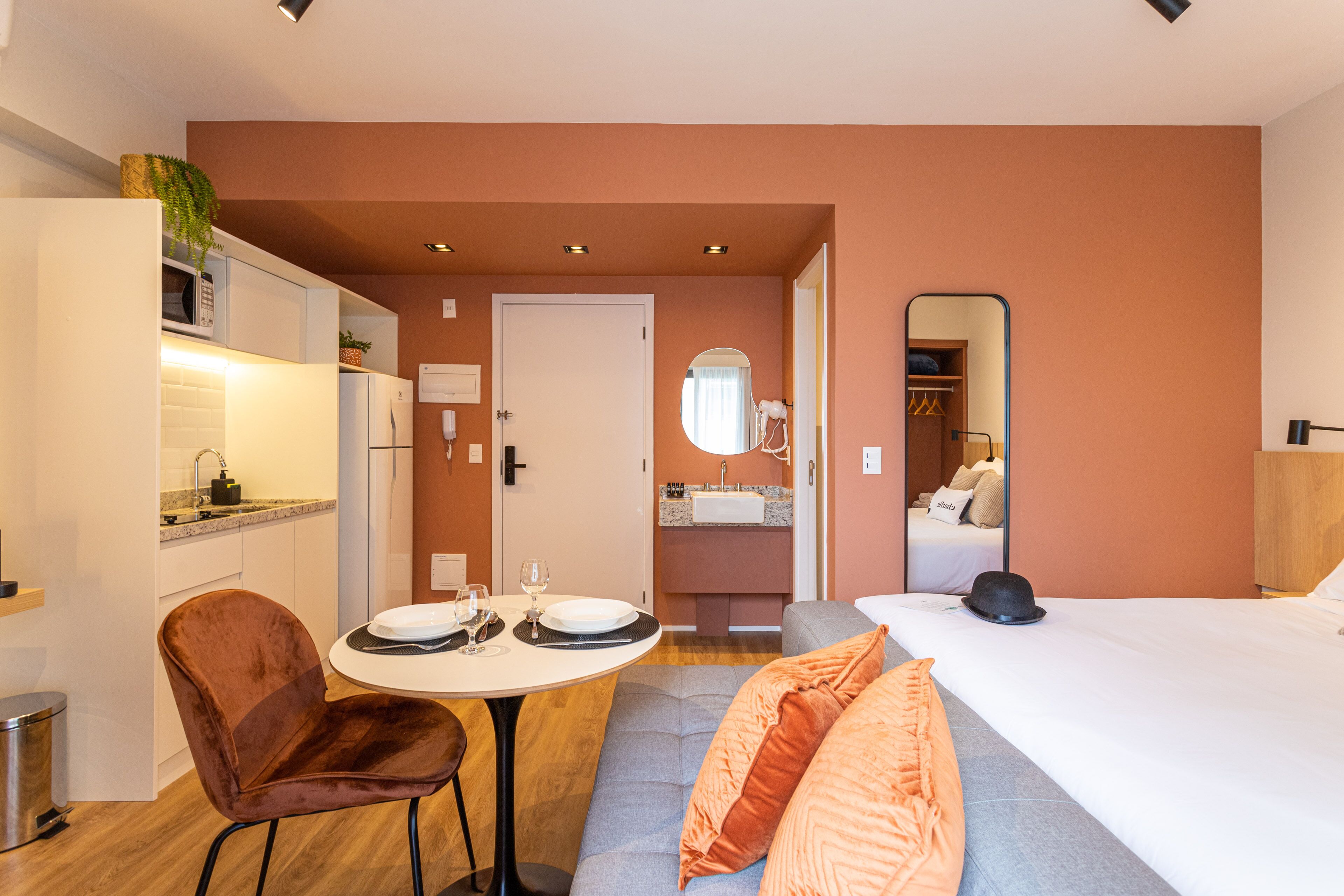 Ein kompaktes Hotelzimmer mit Küchenzeile, einem Essbereich mit rundem Tisch und zwei Stühlen, einer Couch mit orangenen Kissen, einem Bett und einem großen Spiegel an der Wand.