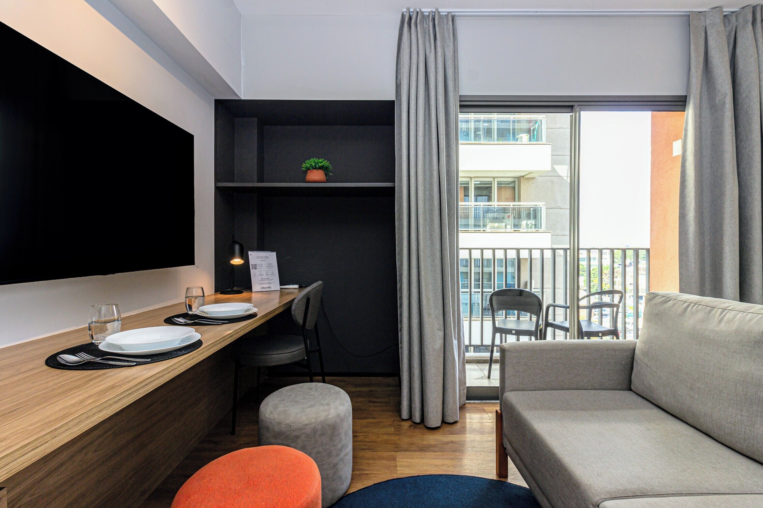 Un salon moderne avec un canapé, une table à manger avec de l’assiette et des couverts, un téléviseur fixé au mur et un balcon avec des chaises.