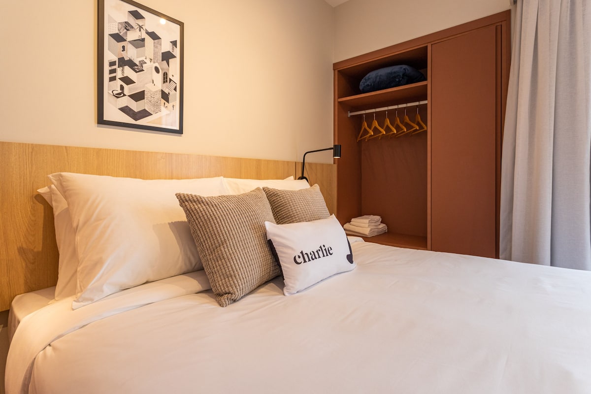 A neatly made bed with white and beige bedding, a framed picture on the wall, a wooden headboard, and a wooden wardrobe with hanging space.