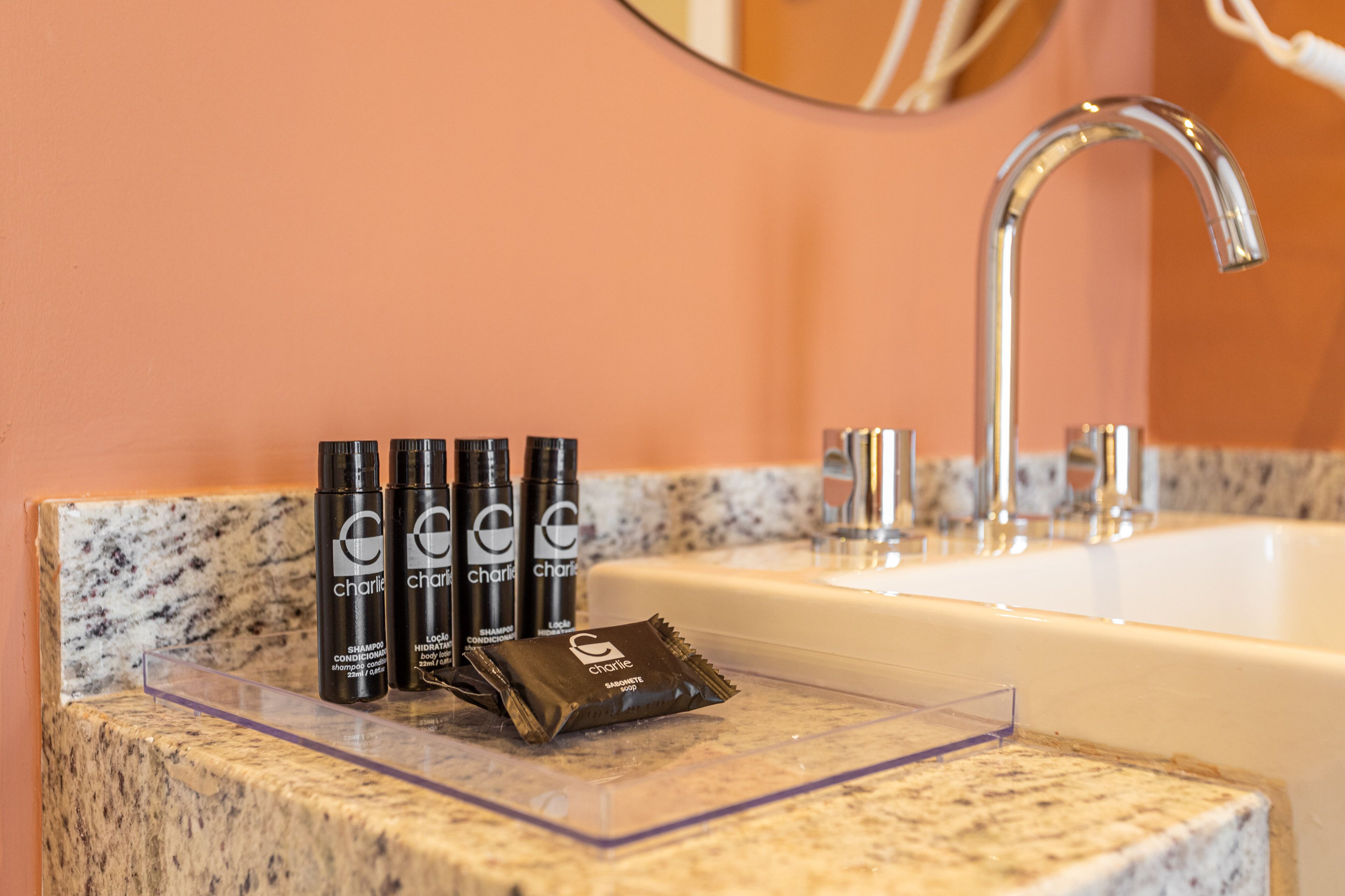 A bathroom sink with a marble countertop, three bottles of Charlie shampoo, and a packet of Charlie body lotion.