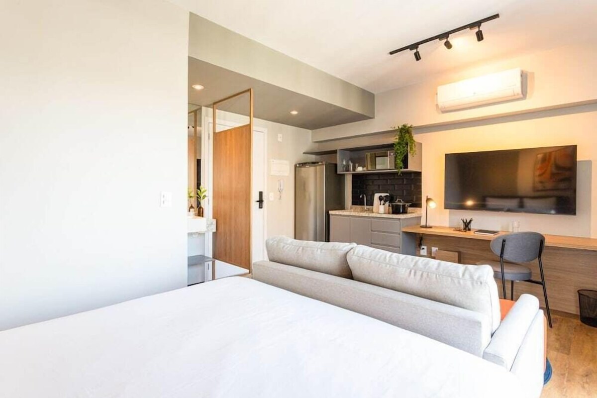 A modern bedroom with a large bed, a desk with a chair, a TV, and a kitchen area in the background.