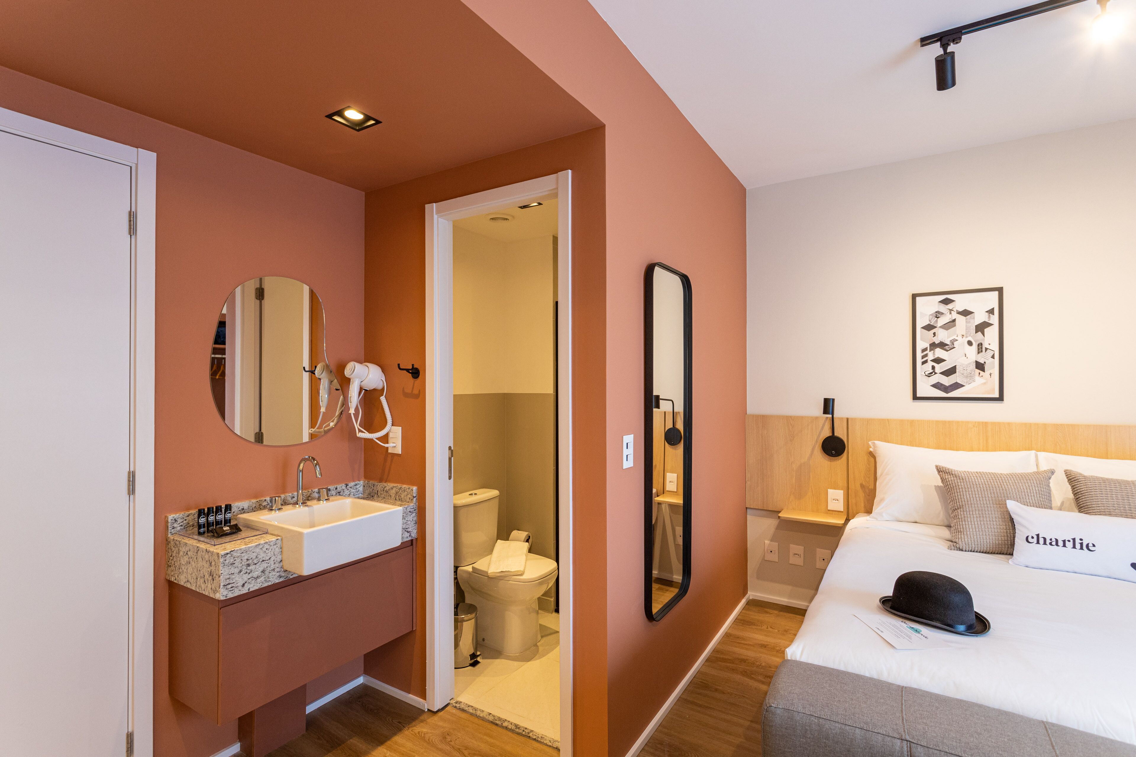 A modern hotel room with a bathroom, a bed with a hat, and a framed picture on the wall.