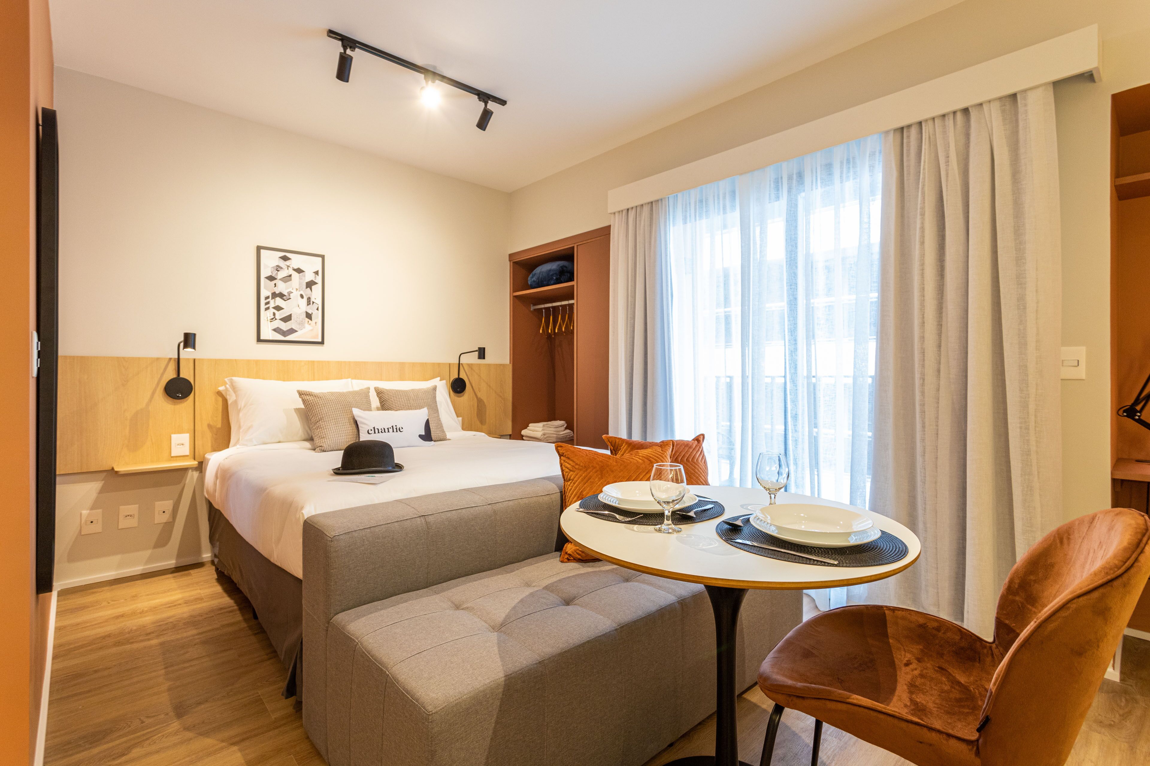 A hotel room with a bed, a dining table with chairs, a bench, a wardrobe, and a window with curtains.