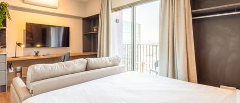 A modern hotel room with a large bed, a flat-screen TV, a desk with a chair, and a balcony with a view of the city.
