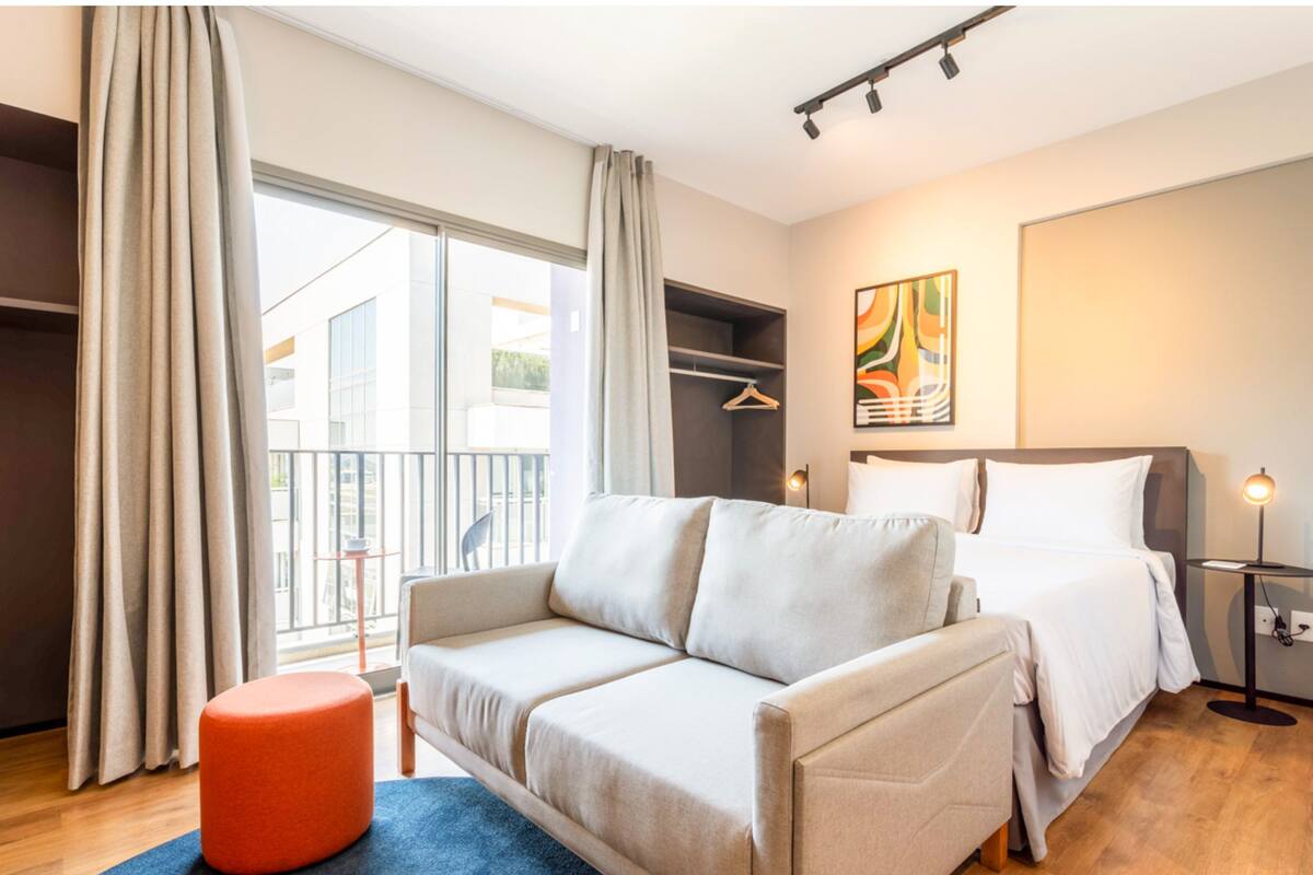 A modern hotel room with a sofa, a bed, a small orange ottoman, a painting on the wall, and a view of the outside balcony.