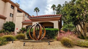 A building with a circular metal sculpture in front, surrounded by landscaped gardens and palm trees. - Biltmore Retreat Resort-Style Heated Pool Spa Gym (Phoenix)
