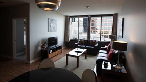 Un salon moderne avec un canapé, une table basse et un téléviseur. Une grande fenêtre offre une vue sur les bâtiments environnants.
