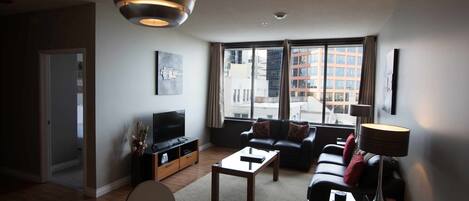 Een moderne woonkamer met een bank, salontafel en televisie. Er is een groot raam met uitzicht op gebouwen.
