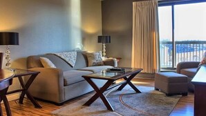 Un salon moderne avec un canapé, une table basse et un fauteuil, de grandes fenêtres avec des rideaux et une vue sur l’extérieur.