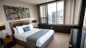 Une chambre d’hôtel avec un grand lit, deux tables de chevet avec des lampes, une vue sur les immeubles par la fenêtre et des œuvres d’art au mur.