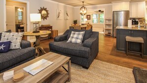Een ruime woonkamer met een bank, salontafel en een eethoek met stoelen. Achtergrond toont een keuken met een koelkast en kastjes.