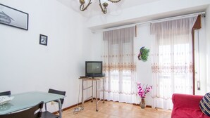 Una sala de estar con una mesa de vidrio, sillas, un televisor sobre un mueble bajo, un sofá rojo y una ventana con cortinas.