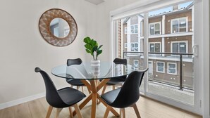 A modern dining area with a glass table, black chairs, a potted plant, and a woven mirror on the wall.