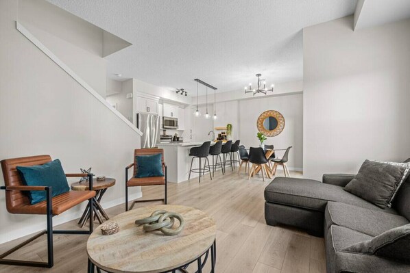 A modern living room with a sofa, chairs, a dining area with a table, and a kitchen in the background. - Fireside Vista | King Bed | Cable | Pets | Modern (Cochrane)