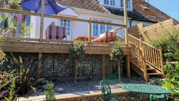 A wooden deck with a stone wall, a wooden staircase, and a green metal table and chair. - Waverley (Lyme Regis)