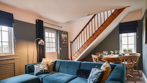 A living room with a blue sofa, a wooden staircase, a dining table with chairs, and a view of the outdoors. - Waverley (Lyme Regis)