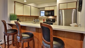 A modern kitchen with a stainless steel refrigerator, a microwave, and a stove. There are three wooden bar stools and a potted plant. - Skiers! Fox Hollow 2br/2ba Condo. Across From Pico, Minutes To Killington 2 Bedroom Condo (Rutland)