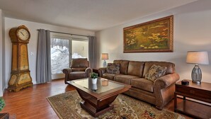 A living room with a brown leather sofa, a glass-top coffee table, a wooden floor, a large painting, and a grandfather clock. - Skiers! Fox Hollow 2br/2ba Condo. Across From Pico, Minutes To Killington 2 Bedroom Condo (Rutland)