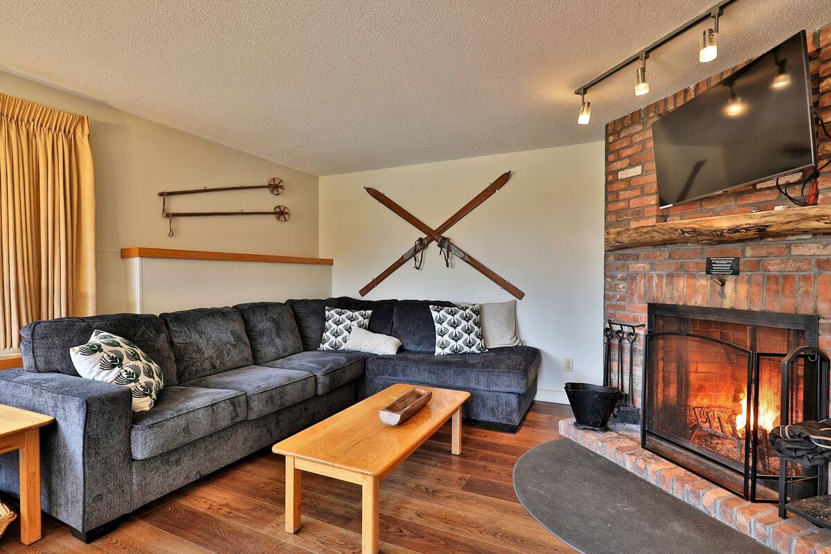 A cozy living room with a sectional sofa, a wooden coffee table, a fireplace, and skis mounted on the wall.