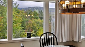 A dining area with a table and chairs, a chandelier, and a view of a forested landscape through large windows. - Fall Line Condos by Killington VR - 2 Bedrooms (Killington)