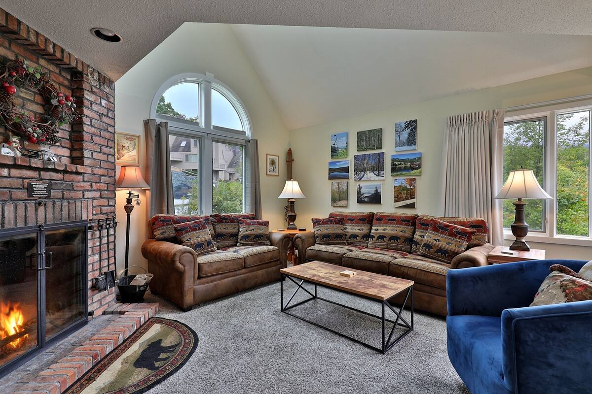 A cozy living room with a fireplace, two sofas, a coffee table, and a large window with curtains.