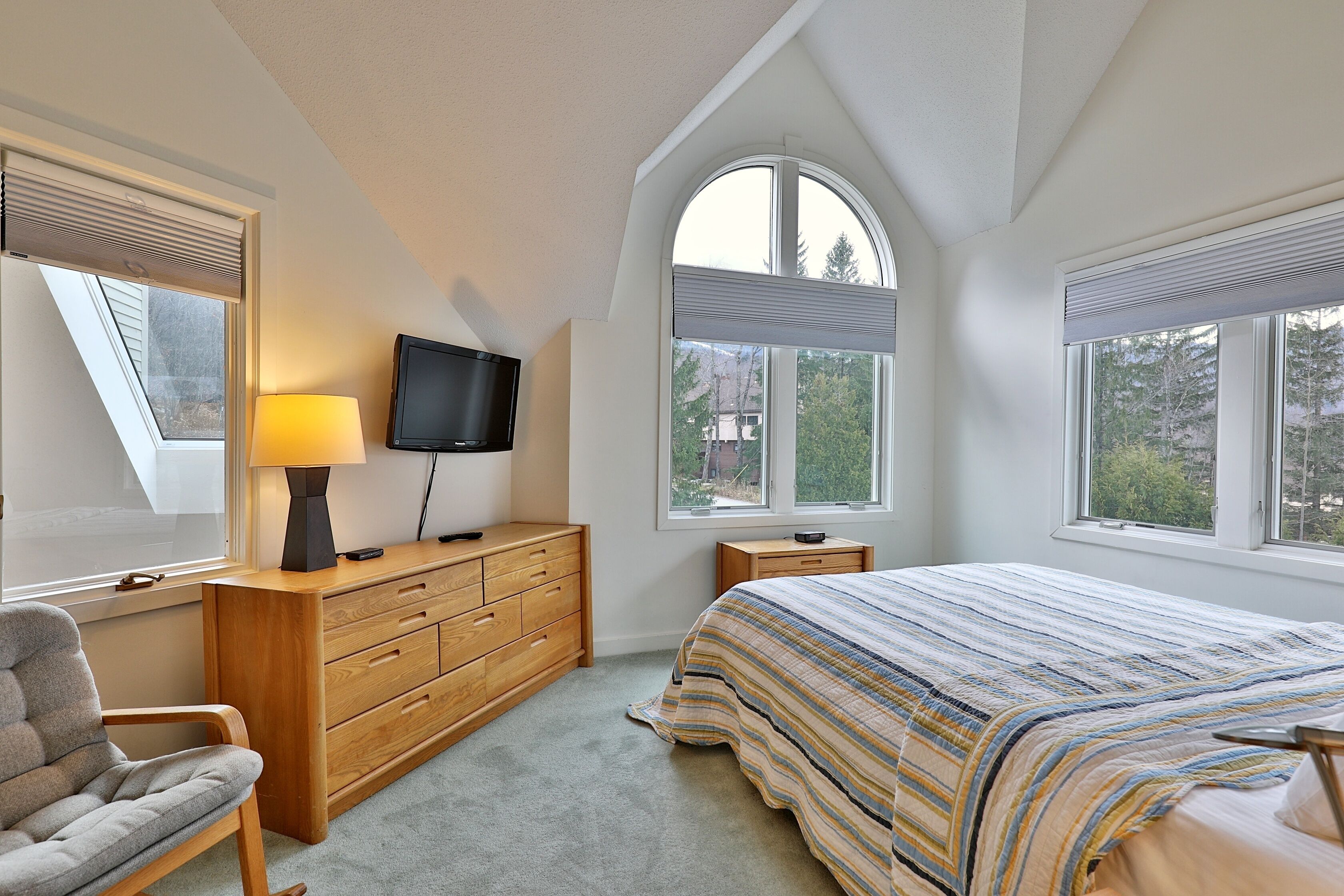 A bedroom with a bed, a dresser, a chair, a TV, and large windows.