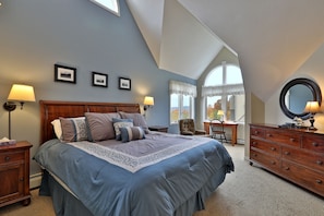 A bedroom with a large bed, a dresser, a chair, and a desk. There are framed pictures on the wall and a window with curtains.