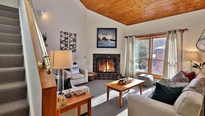 A cozy living room with a fireplace, a staircase, and a large window with curtains. - Trail Creek by Killington Vacation Rentals (Killington)