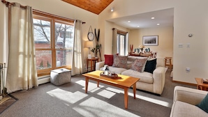 A spacious living room with a wooden ceiling, large windows with curtains, a sofa, a coffee table, and a fireplace. - Trail Creek by Killington Vacation Rentals (Killington)