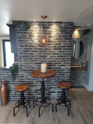 Un coin repas avec un mur en briques, deux tabourets en bois, une table ronde, un luminaire suspendu et une bougie.
