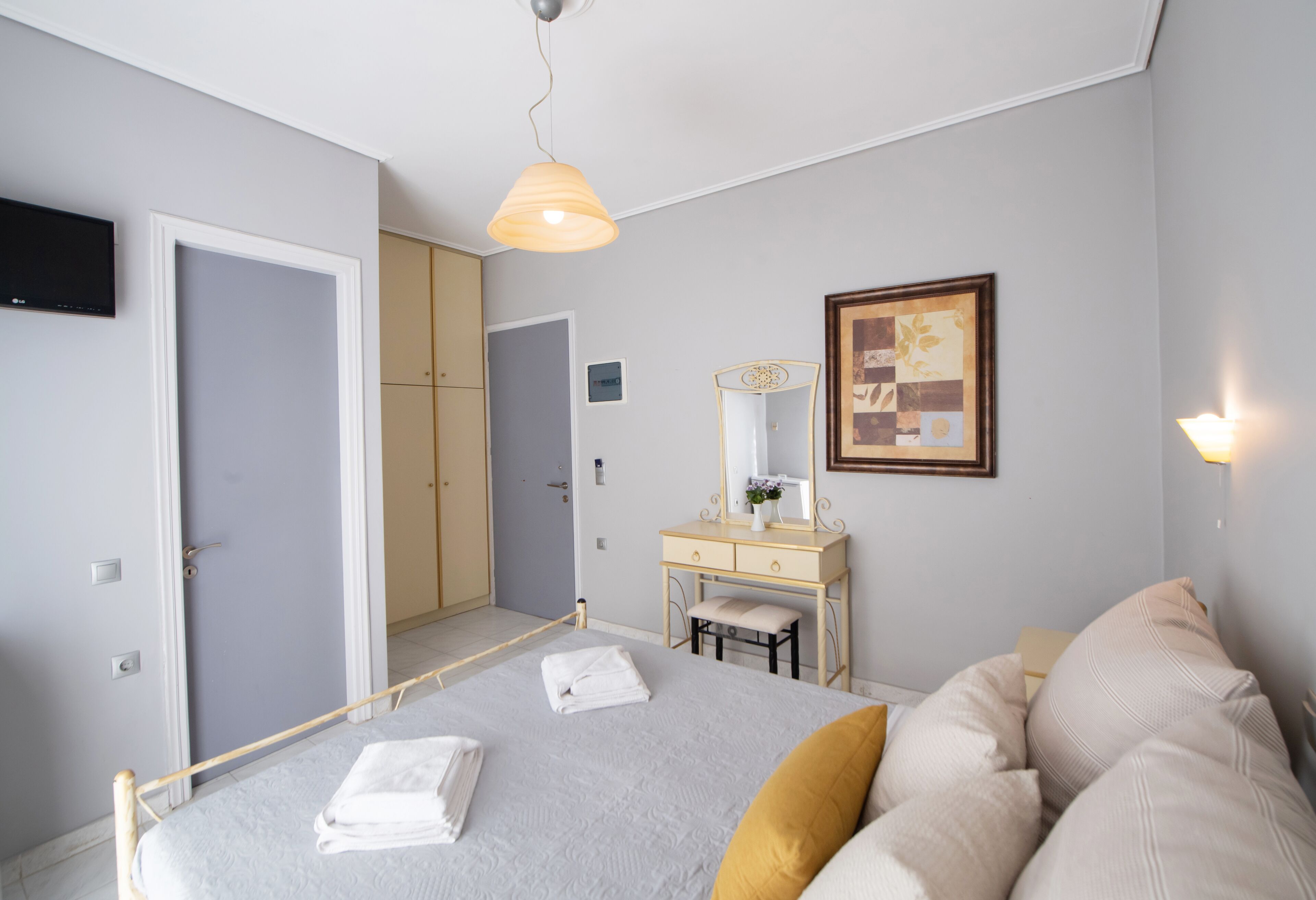 Ein Schlafzimmer mit Bett, Nachttisch und Spiegel. Eine Tür und ein an der Wand befestigtes Fernsehgerät sind ebenfalls vorhanden.