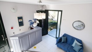 A modern living room with a blue sofa, a kitchen area with a white cabinet, and a large window offering a view of the outdoors. - George House (Lefkada)
