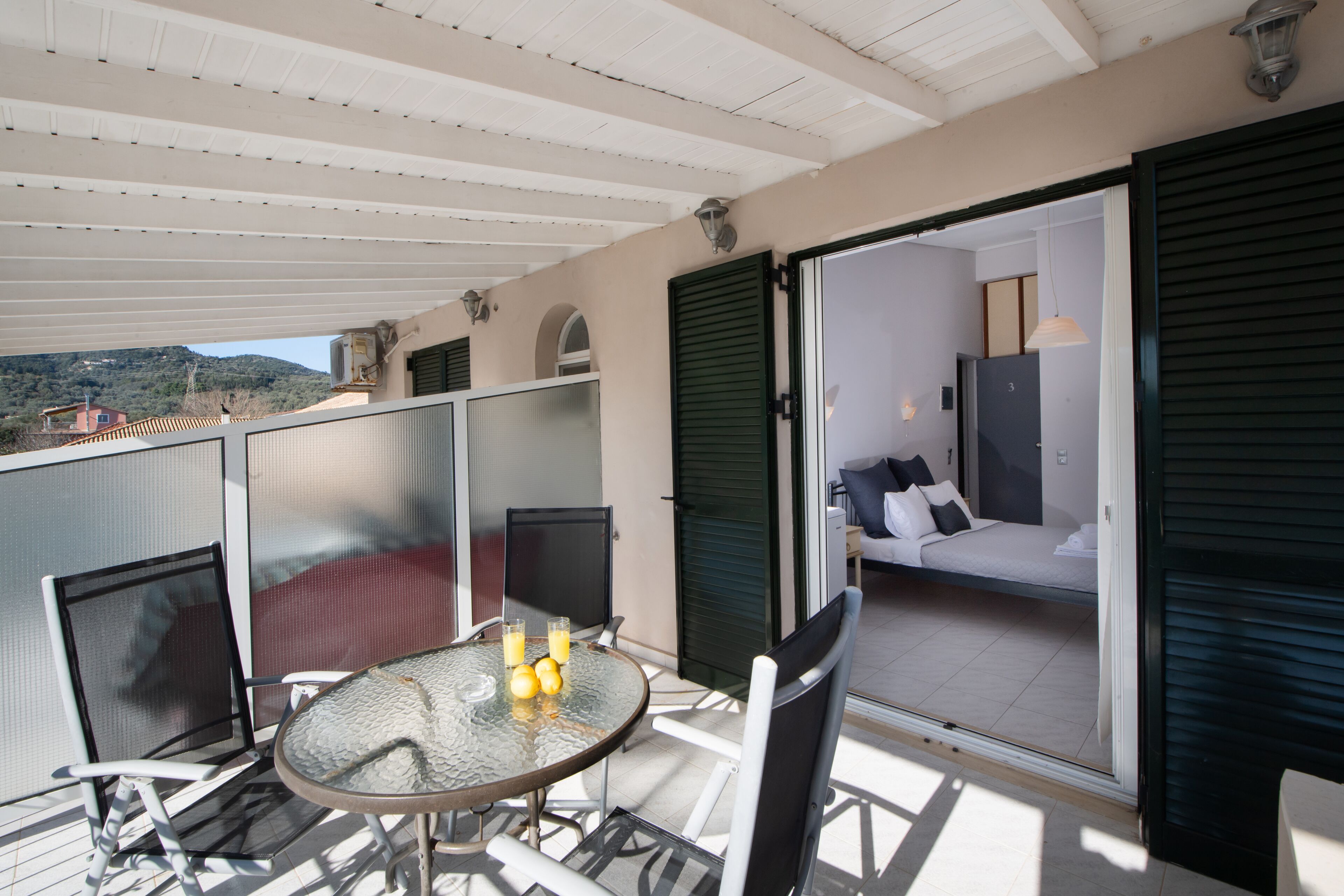 A balcony with a glass table and chairs, a bed with pillows, and a view of the hills.