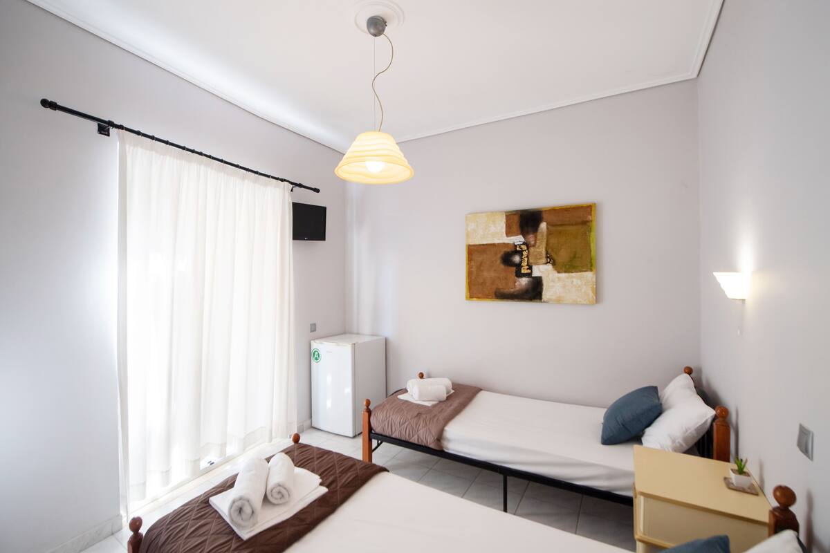 Una habitación de hotel con dos camas, un escritorio pequeño, un calentador, una ventana con cortinas y un cuadro en la pared.