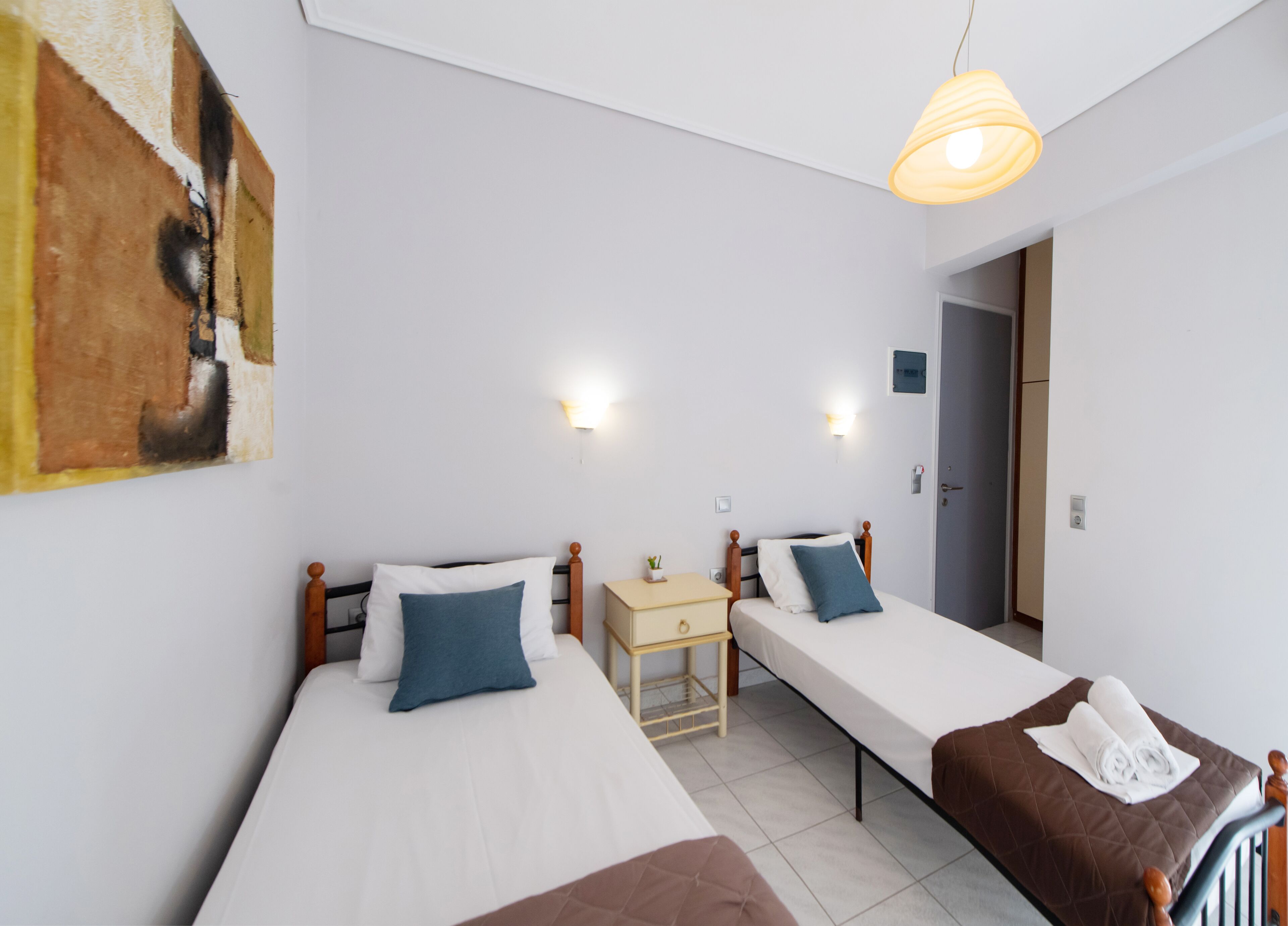 A room with two single beds, a small bedside table with a lamp, a painting on the wall, and a door leading to another room.