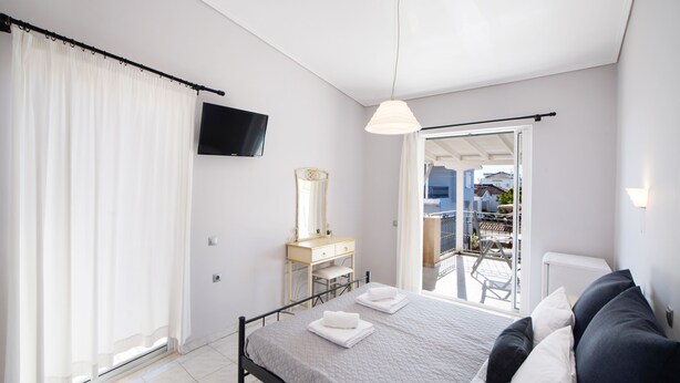 A bedroom with a bed, a dresser, a mirror, a TV, and a balcony with a view of the city. - George House (Lefkada)