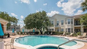 A swimming pool with lounge chairs and umbrellas, surrounded by multi-story buildings with balconies. - Legacy Villa 1901 | Just a Short Walk to the Beach! (Gulfport)