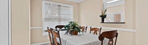 A dining area with a table set for four, surrounded by wooden chairs. A large potted plant is in the center of the table.