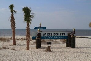 A welcome sign at the Mississippi Gulf Coast beach with palm trees and the ocean in the background.