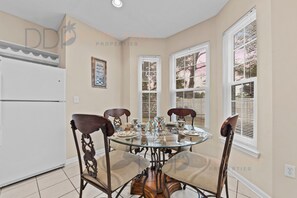 A dining area with a glass table, four chairs, a refrigerator, and windows with blinds.