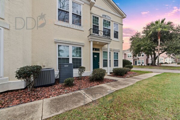 A two-story residential building with a front porch, a small garden with shrubs, and an air conditioning unit.