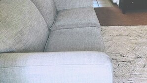A living room with a sofa, a coffee table, and a television. There is a window with blinds, a refrigerator, and a chair in the background.