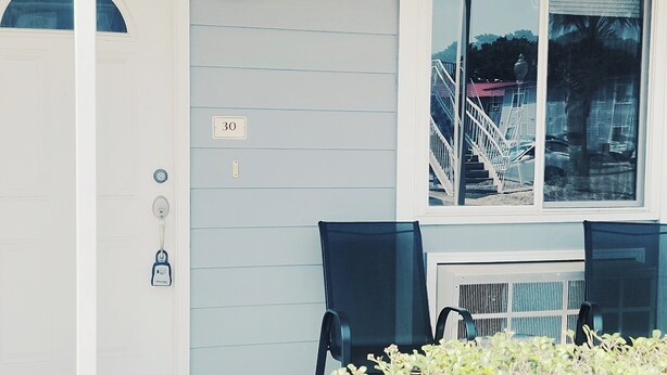 A house exterior with a number 30 sign, a white door, a window with a view of a staircase and palm tree, and a bench outside.