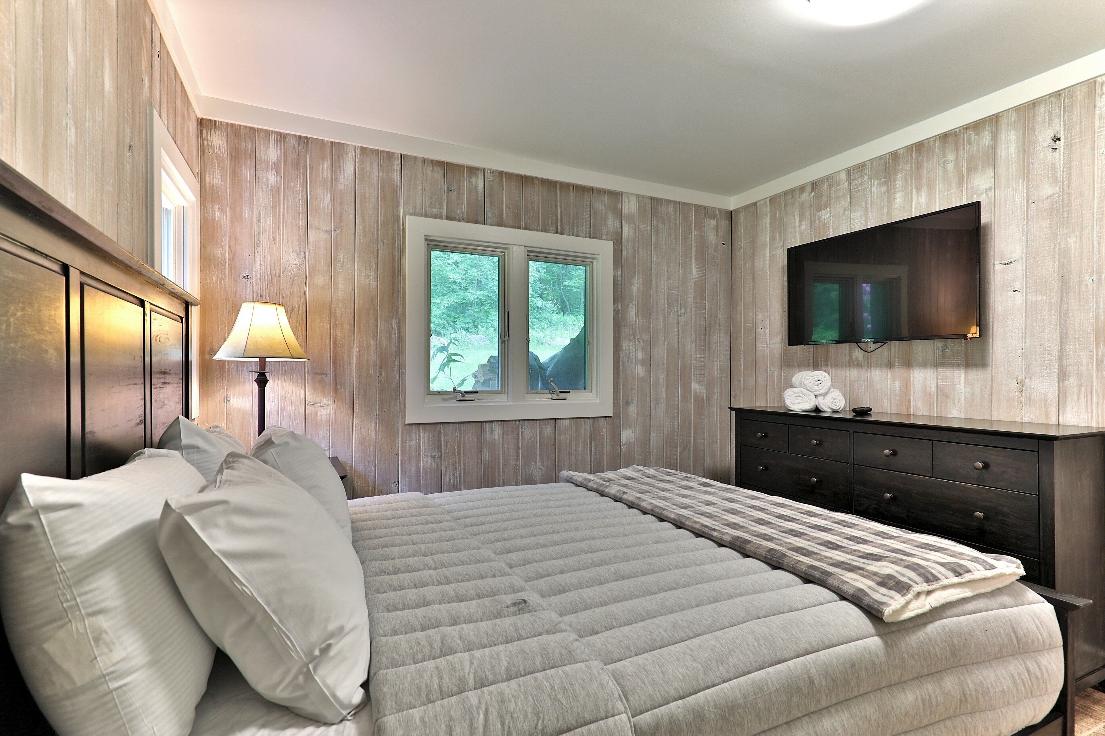 A bedroom with a large bed, a dresser, a TV mounted on the wall, and a window with a view of trees.
