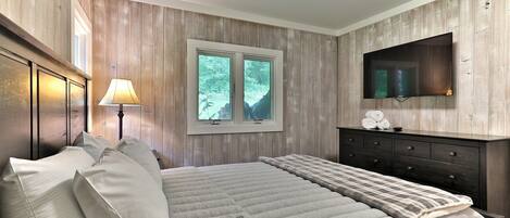 A bedroom with a large bed, a dresser, a TV mounted on the wall, and a window with a view of trees.